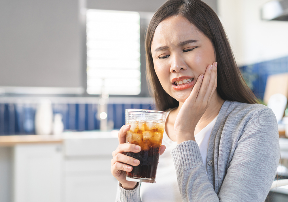 A woman is holding her jaw with one hand and sipping a soda with the other.